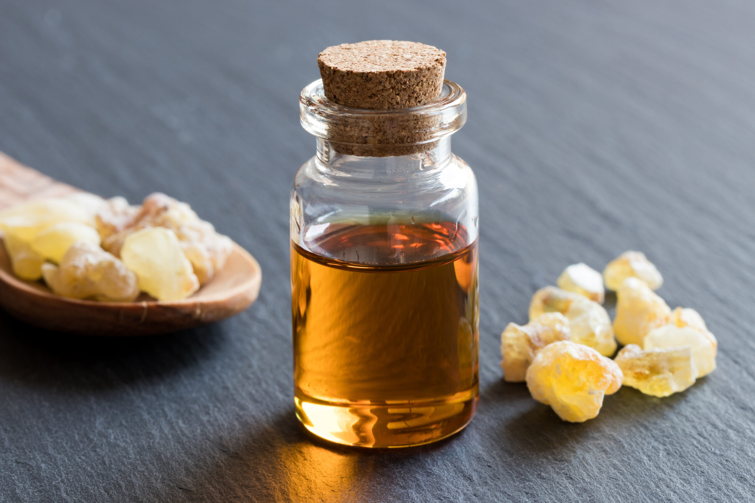 Glass jar of frankincense oil with cork stopper, surrounded by frankincense resin pieces on a dark slate background, highlighting natural skincare ingredients for Yeshua Gold products.