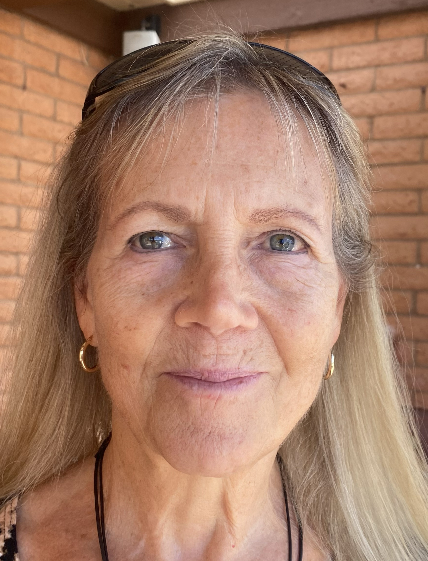Woman with long blonde hair and earrings, showcasing healthy skin and a natural glow, standing against a brick wall, reflecting positive effects of Yeshua Gold skincare products.